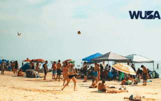 Crowded Beach Trip scene with people relaxing, playing volleyball, and sitting under umbrellas and tents. WUSA logo in corner.