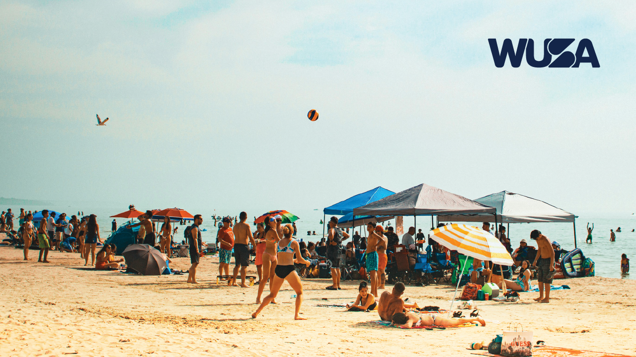 Crowded Beach Trip scene with people relaxing, playing volleyball, and sitting under umbrellas and tents. WUSA logo in corner.