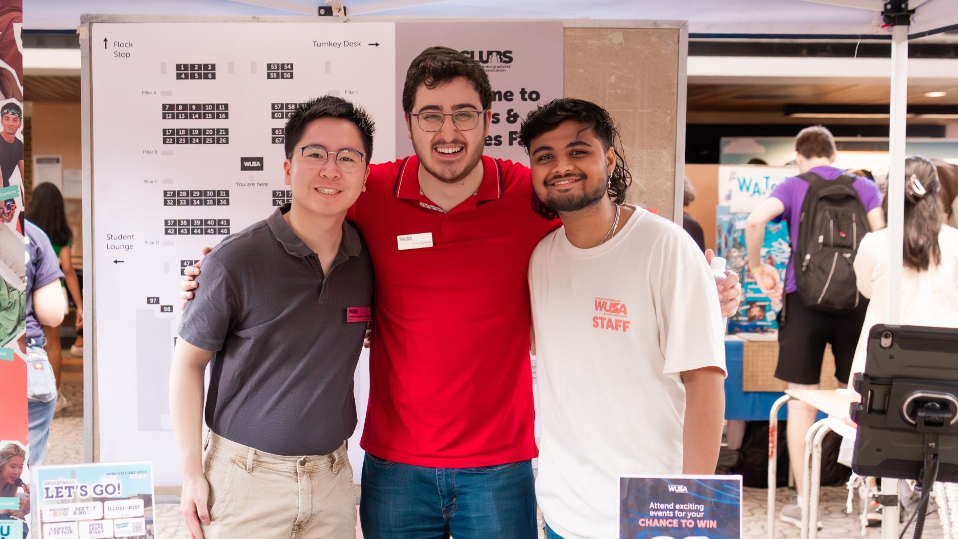 Three smiling young men stand together at an event booth, posing for a photo in front of informational posters highlighting student groups and the student experience.
