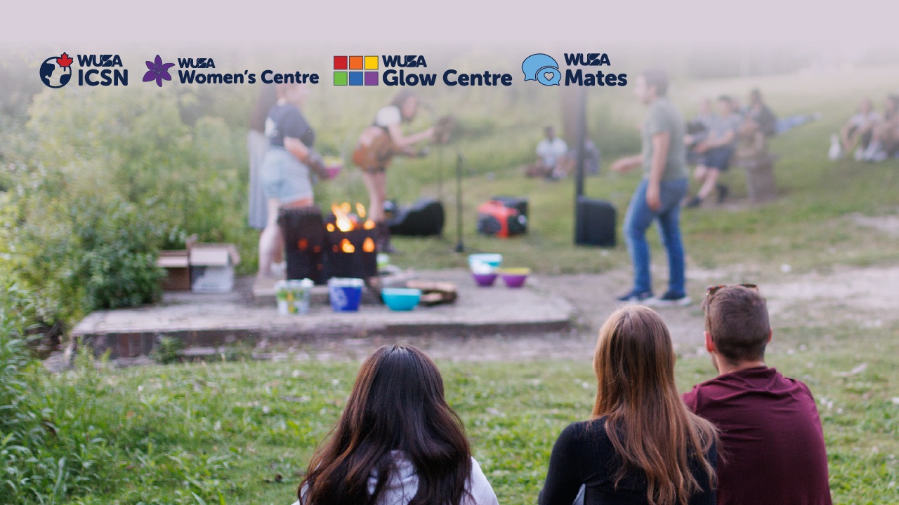 Three people sit on grass watching a group gather around a campfire, celebrating Bonfire Night; four WUSA centre logos appear above.
