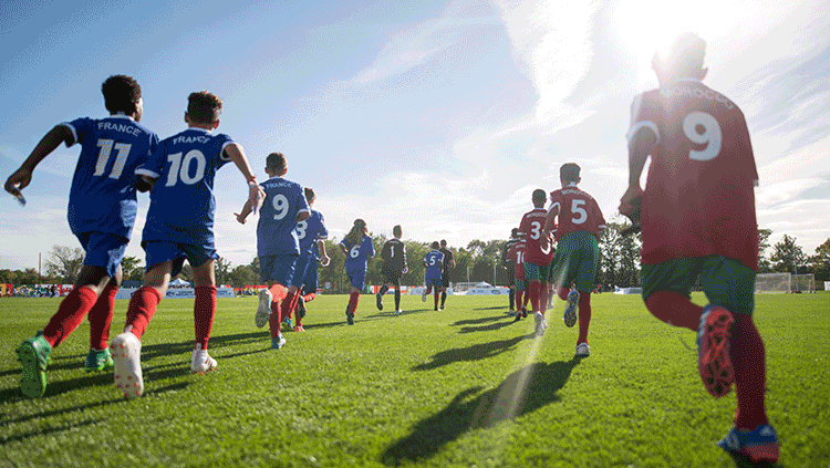 Young soccer players in blue and red uniforms run toward the field under bright sunlight, ready to compete in the soccer tournament.