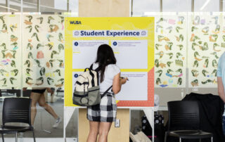 A student with a backpack writes on a "Student Experience" board in a bright, modern space.