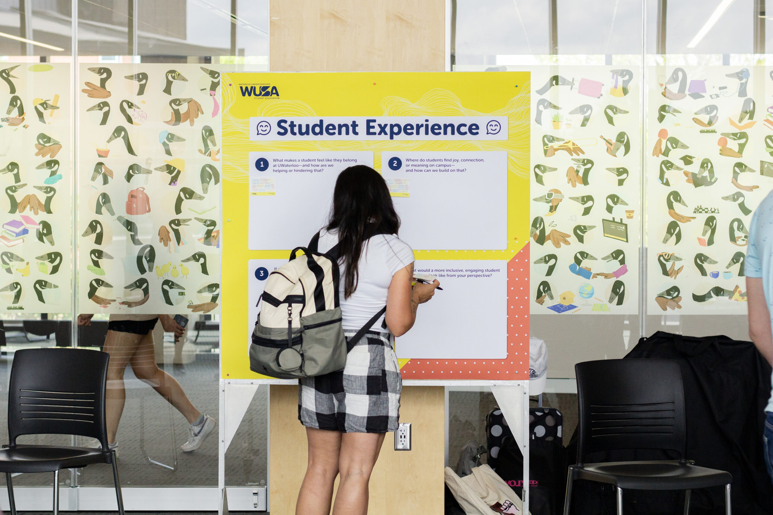 A student with a backpack writes on a "Student Experience" board in a bright, modern space.