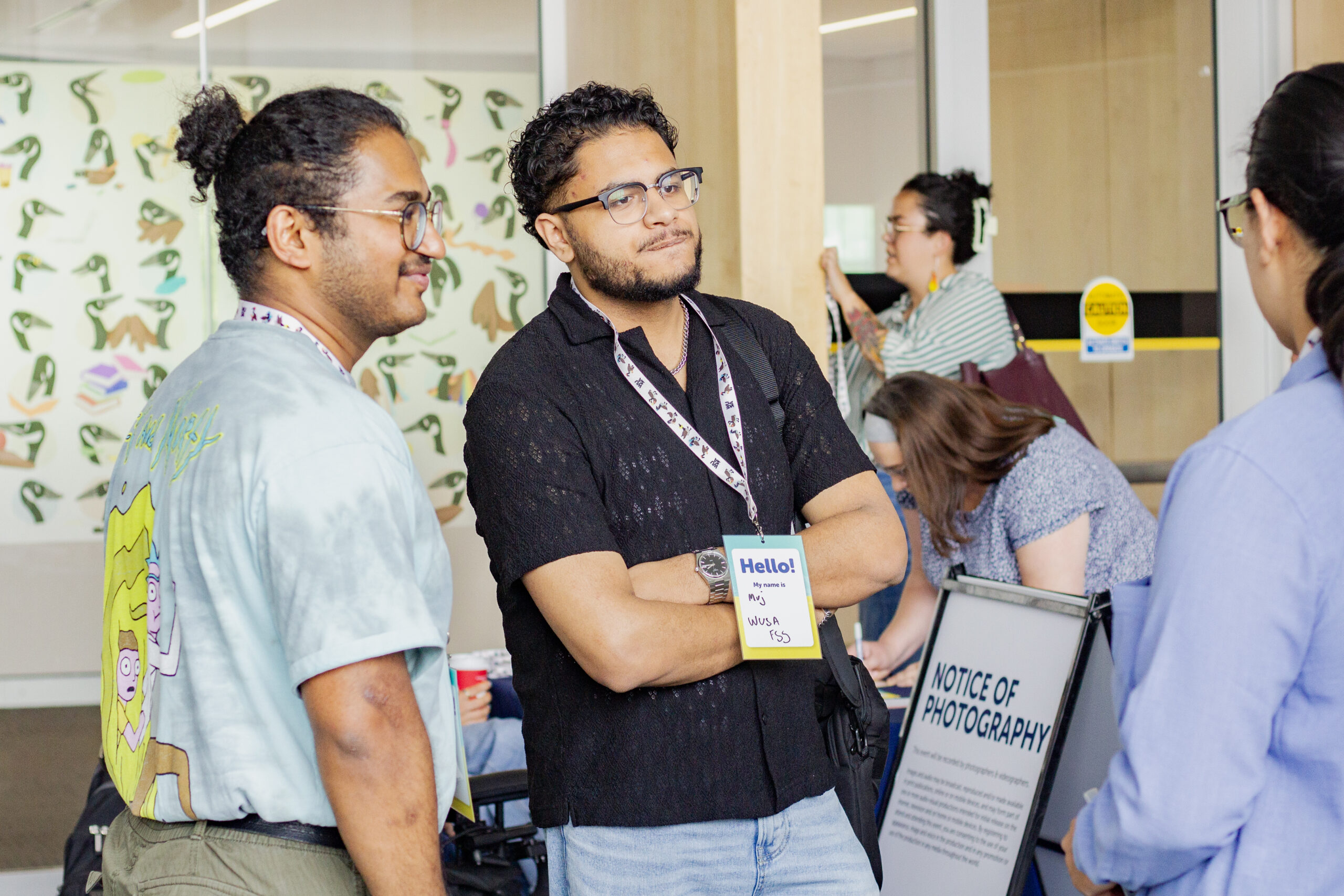 Two people with name tags talk to others at an indoor event near a “Notice of Photography” sign.