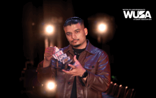 Illusionist Savio Joseph, in a brown jacket, performs a card trick under bright lights with the WUSA logo illuminating the Humanities Theatre backdrop.