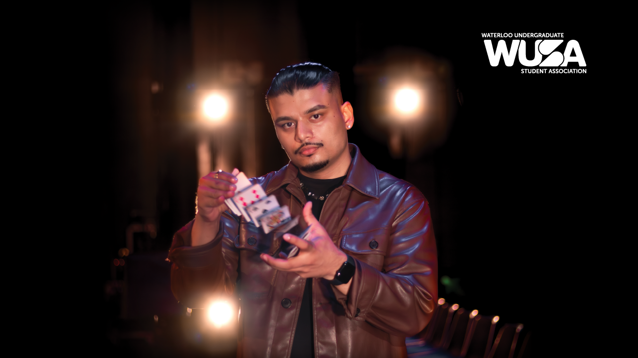 Illusionist Savio Joseph, in a brown jacket, performs a card trick under bright lights with the WUSA logo illuminating the Humanities Theatre backdrop.