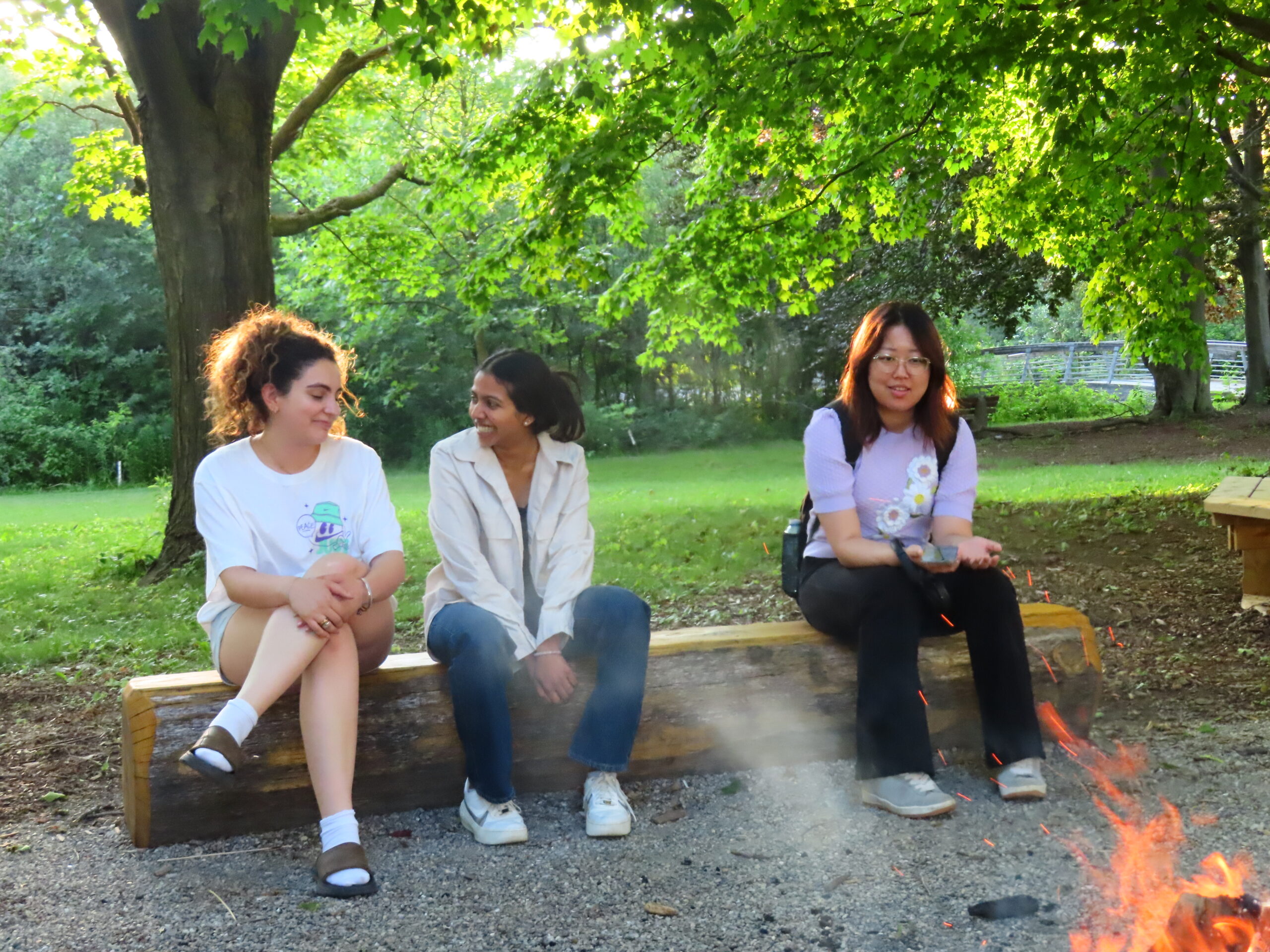 Three young women sit and talk by a campfire in a wooded park on a sunny day, sharing stories of inclusion and equity in their community.
