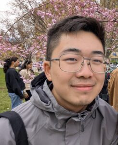 A young man in glasses smiles outdoors under cherry blossoms, celebrating Student Excellence in Spring 2025, with others and trees in the background.