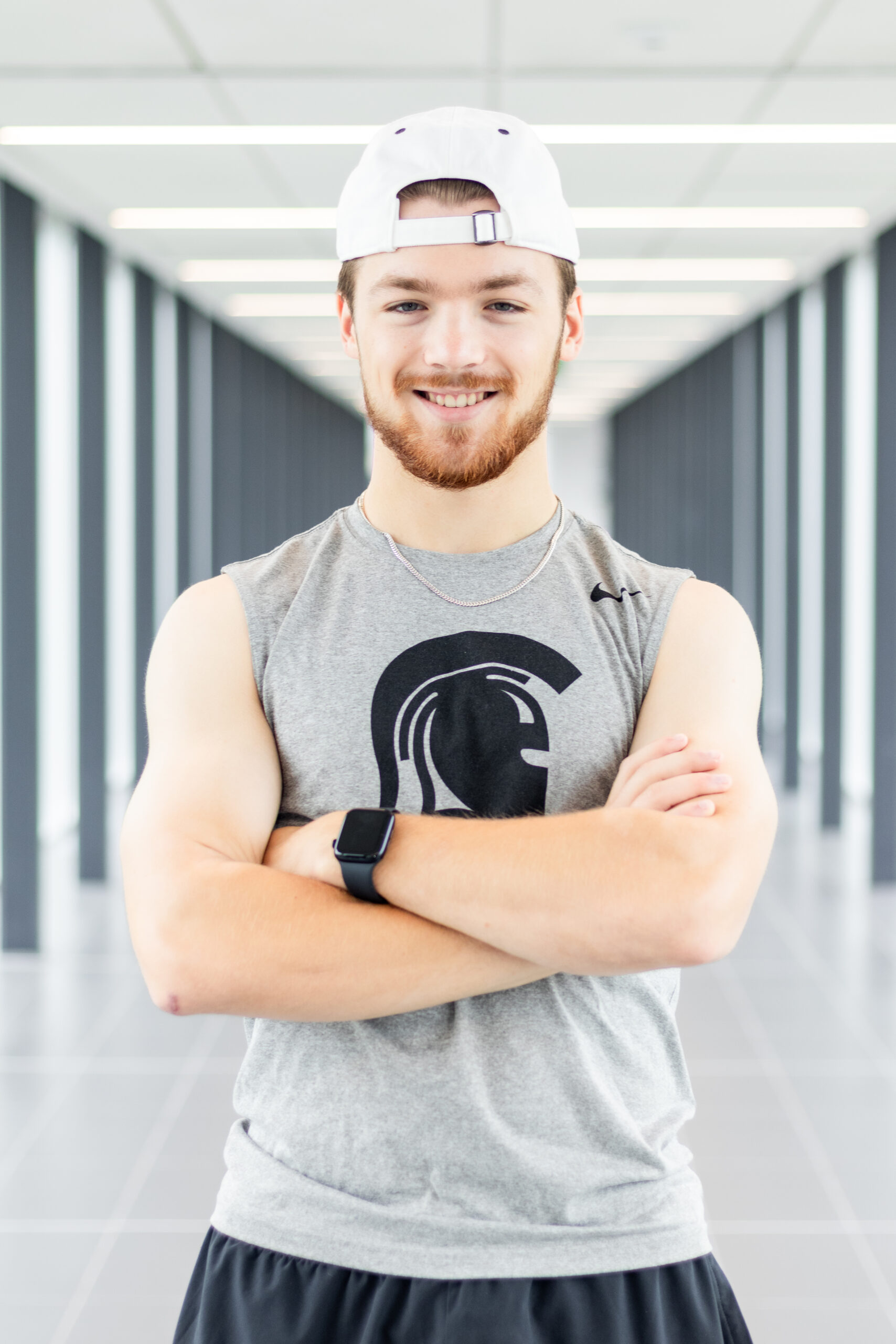 Young man in a sleeveless shirt and white cap stands with arms crossed, smiling in a bright hallway.