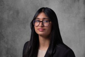 Woman with long dark hair and glasses wearing a black blazer, posing against a gray textured background, honored for Student Excellence at the Spring 2025 WUSA Volunteer Awards.