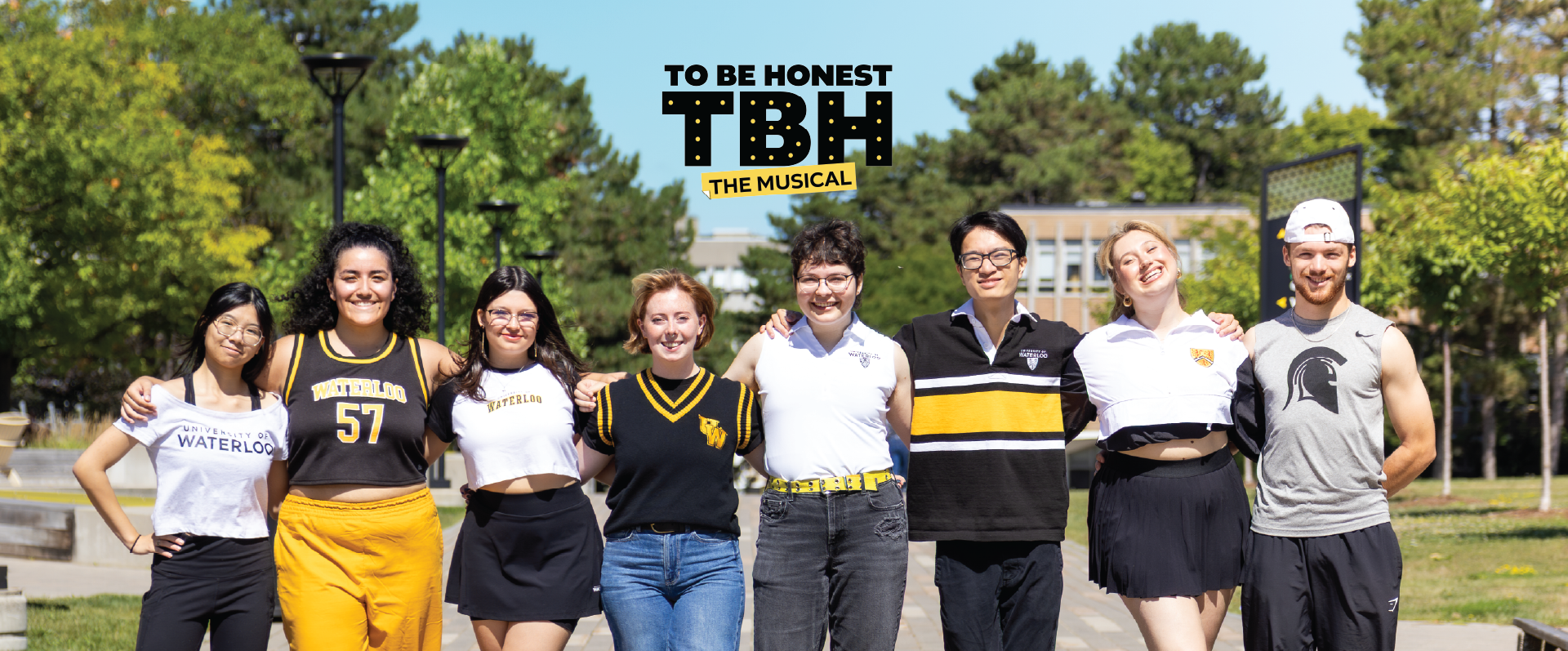 Eight smiling young adults stand arm-in-arm outdoors under a sign for To Be Honest: The Musical, radiating excitement and unity.