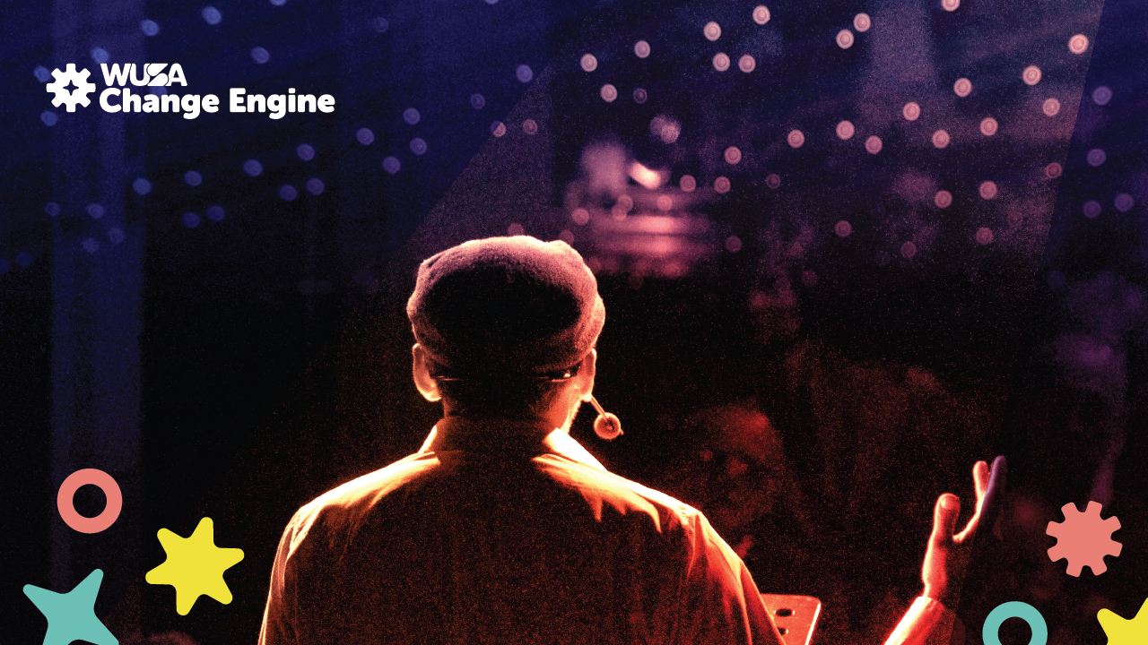 A person speaks on stage to an audience during Live Competition Night, with the WUSA Change Engine logo displayed in the top left corner.