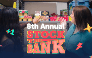 Two women stock shelves with food for the 8th Annual Stock the Bank event, surrounded by colorful Stock the Bank graphics.