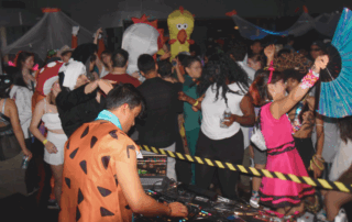 People in colorful Halloween costumes dance at a lively party, with a WUSA DJ playing music in the foreground.