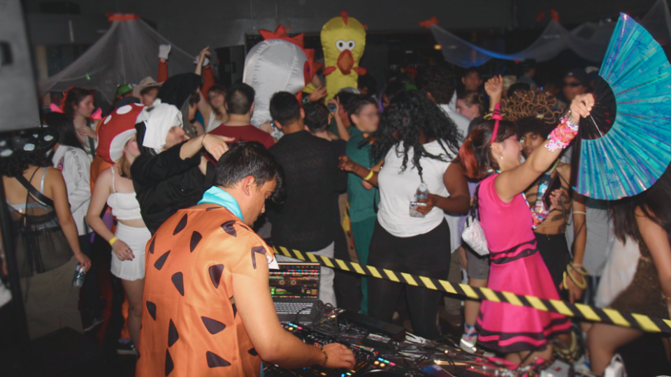 People in colorful Halloween costumes dance at a lively party, with a WUSA DJ playing music in the foreground.