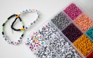 Colorful beads and alphabet beads in a box, with two finished letter bead bracelets reading "BE KIND" and "JUST H"—perfect for your next Bracelet Making night or to inspire creativity at a Weekly Vibes Night.