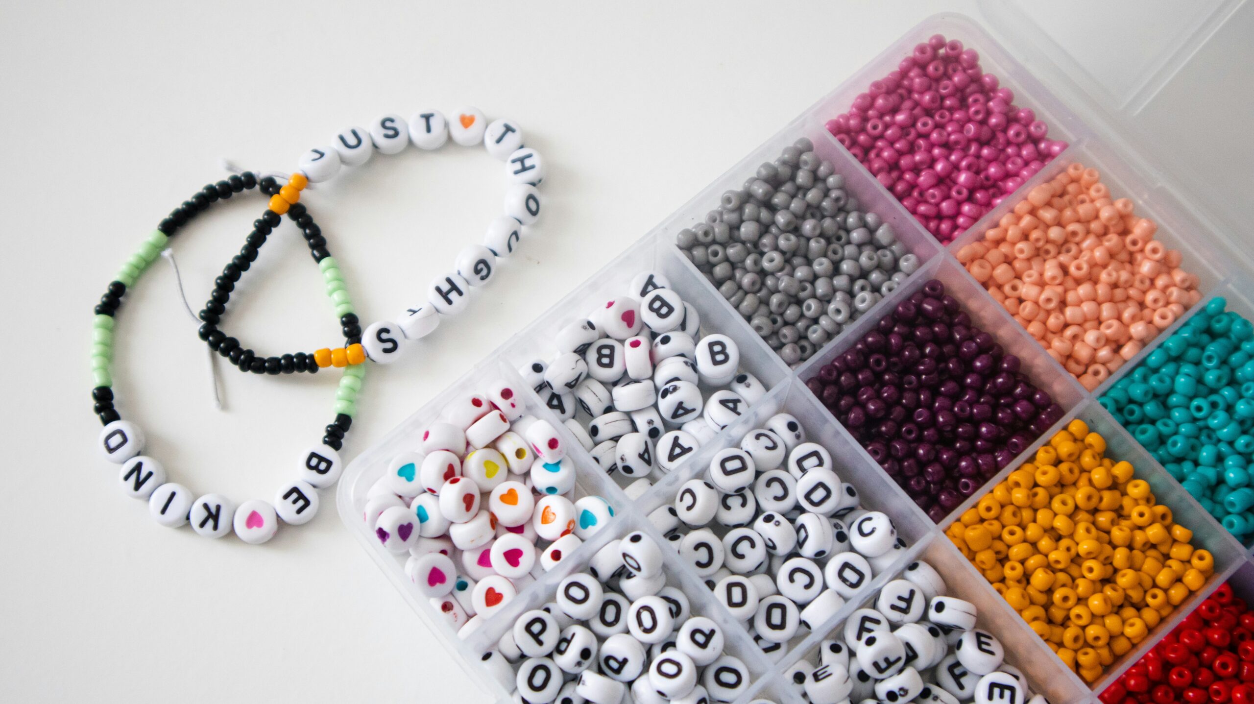 Bracelet Making Colorful beads and alphabet beads in a box, with two finished letter bead bracelets reading "BE KIND" and "JUST H"—perfect for your next Bracelet Making night or to inspire creativity at a Weekly Vibes Night.