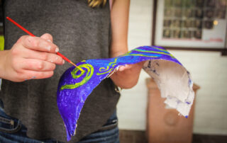 A participant at the Women’s Centre Chest Casting Workshop paints a blue and green design on a papier-mâché mask with a red paintbrush.