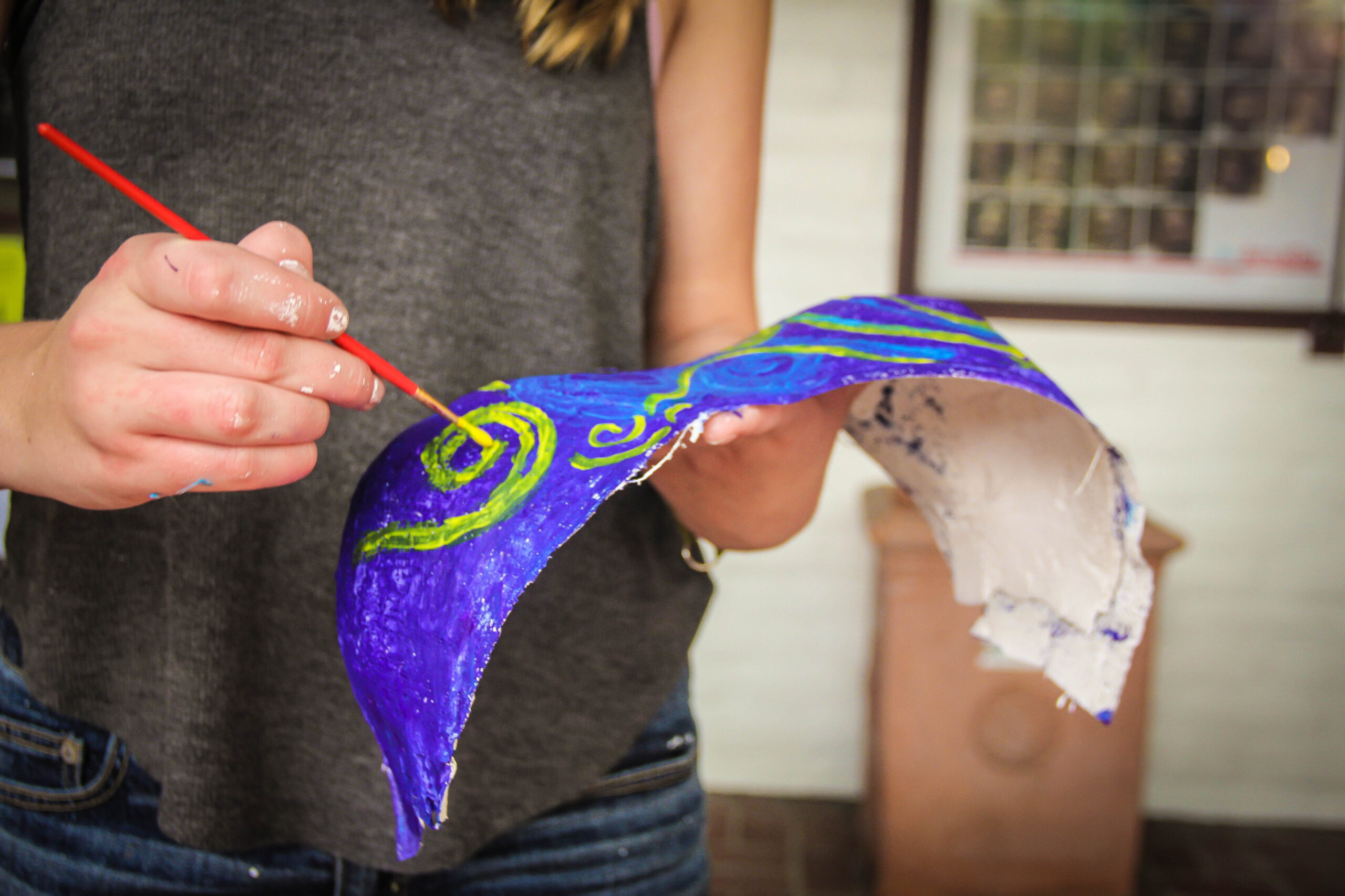 A participant at the Women’s Centre Chest Casting Workshop paints a blue and green design on a papier-mâché mask with a red paintbrush.