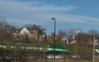 A green and white Go Service commuter train passes by houses and parked cars under a blue sky with wispy clouds, offering a convenient option for commuting to UW.
