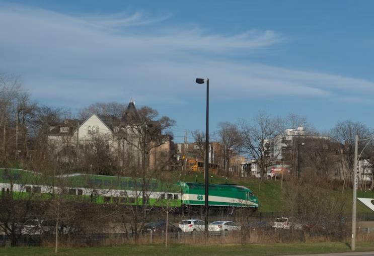 A green and white Go Service commuter train passes by houses and parked cars under a blue sky with wispy clouds, offering a convenient option for commuting to UW.