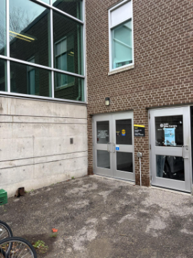 Two gray double doors with signs on a brick building beside a concrete wall and bike racks welcome you as you visit SLC.