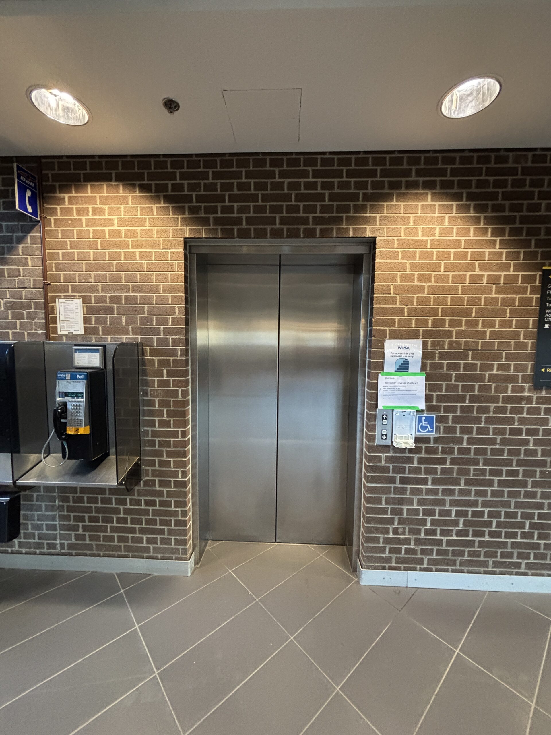 Elevator doors in a brick wall next to a payphone and accessibility buttons, capturing an urban SLC vibe.