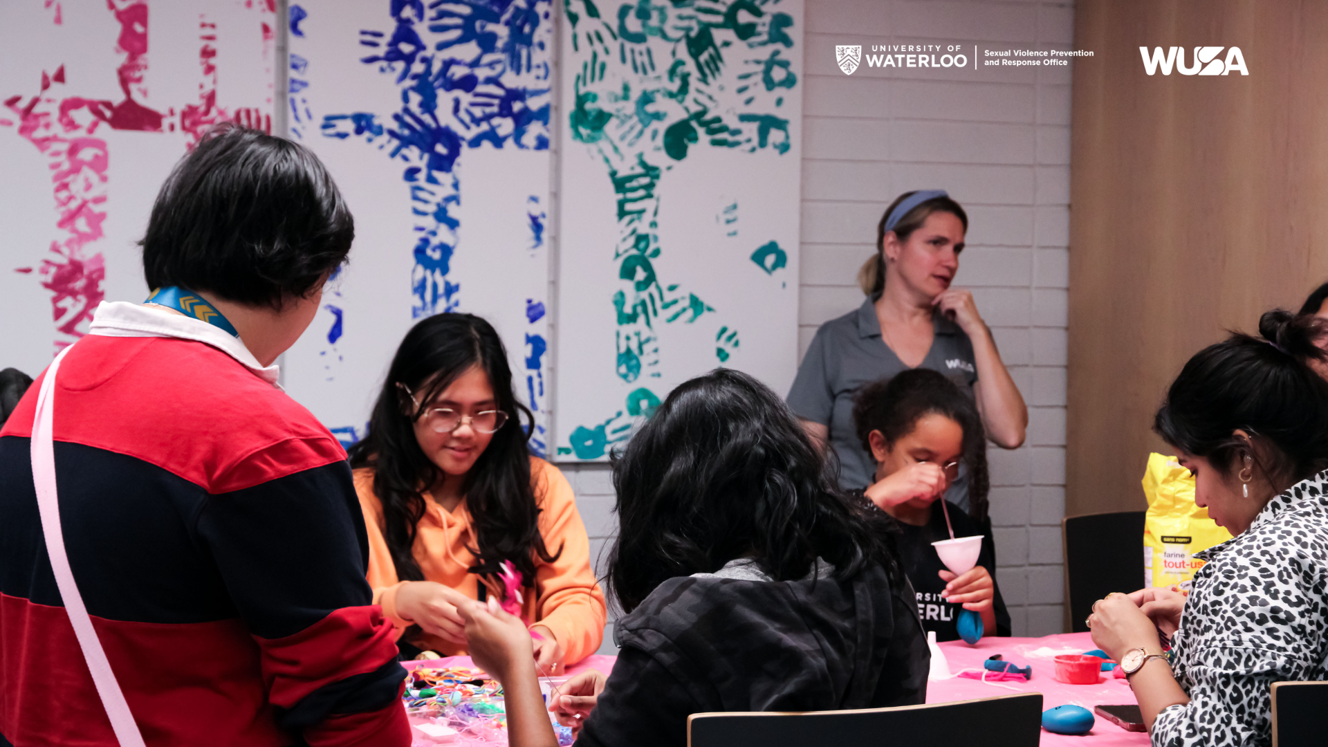 People enjoy a craft session at a table, supervised by an adult, in a colorful room adorned with University of Waterloo logos—a creative escape for all ages.