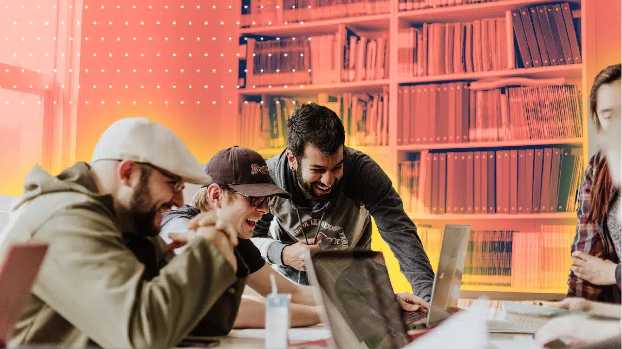 Four people laughing and working together at laptops in a colorful, modern library or office space, planning for Fall 2025 and chatting excitedly about the upcoming WAGS Hockey Night.