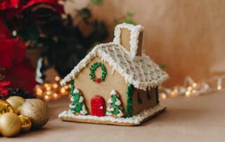A decorated gingerbread house with icing, trees, and a wreath stands ready for the OCC Grand Gingerbread Competition, surrounded by ornaments and festive lights.