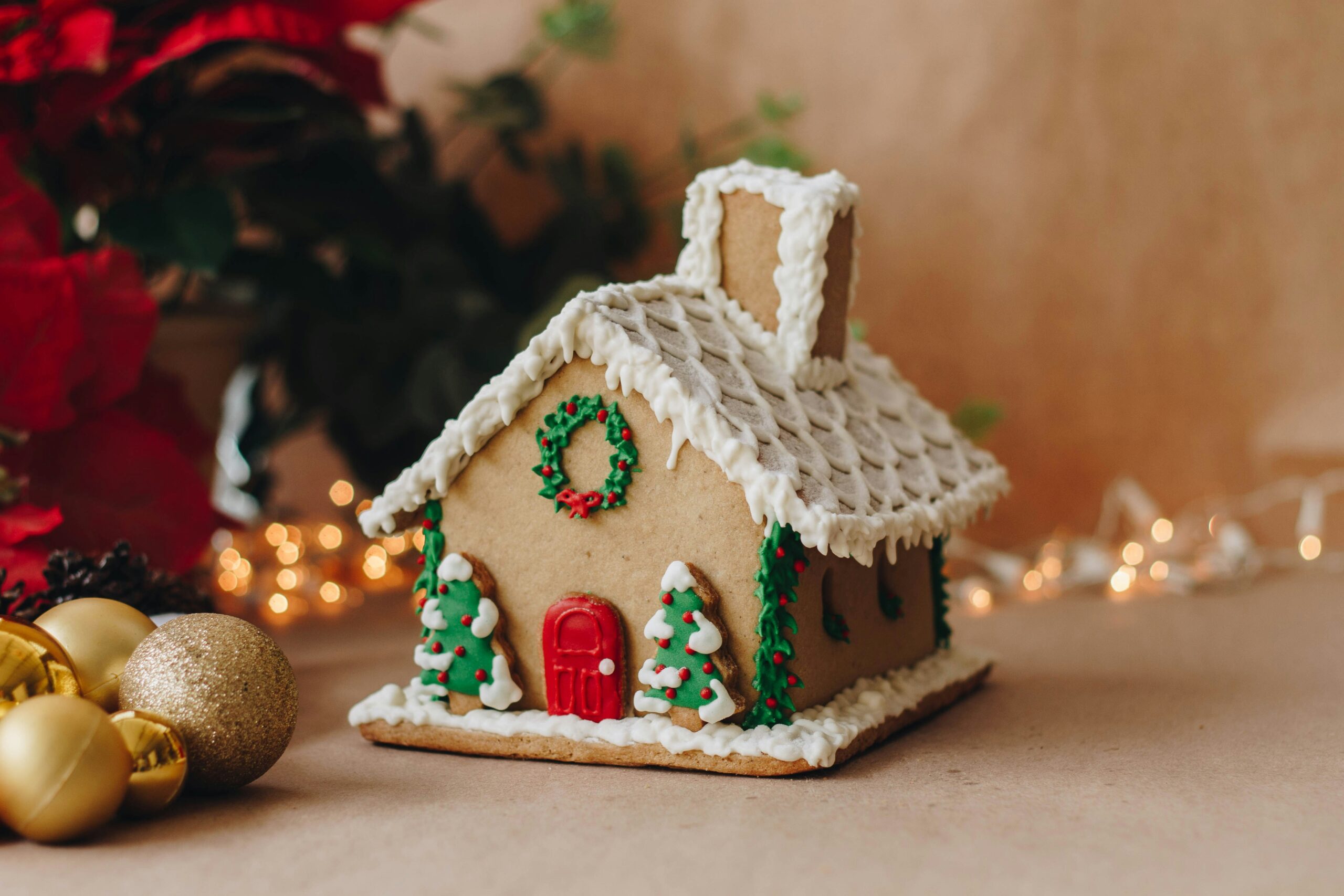 A decorated gingerbread house with icing, trees, and a wreath stands ready for the OCC Grand Gingerbread Competition, surrounded by ornaments and festive lights.