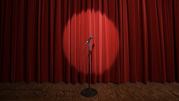 A microphone stands in a spotlight on a wooden floor, ready for Stand Up Comedy, in front of closed red stage curtains.