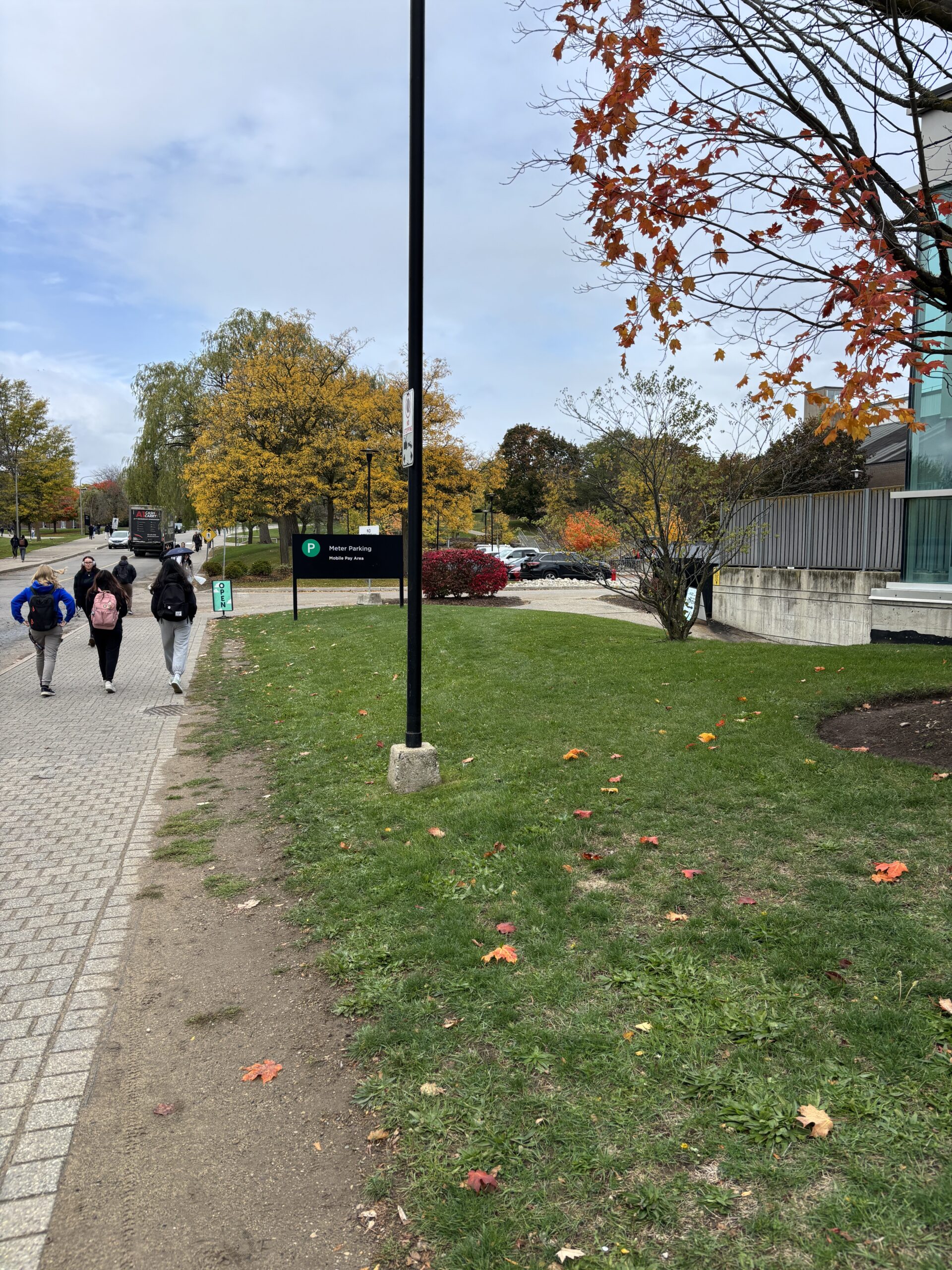 Three people walk on a path near autumn trees and a building on a partly cloudy day, enjoying their visit to SLC.