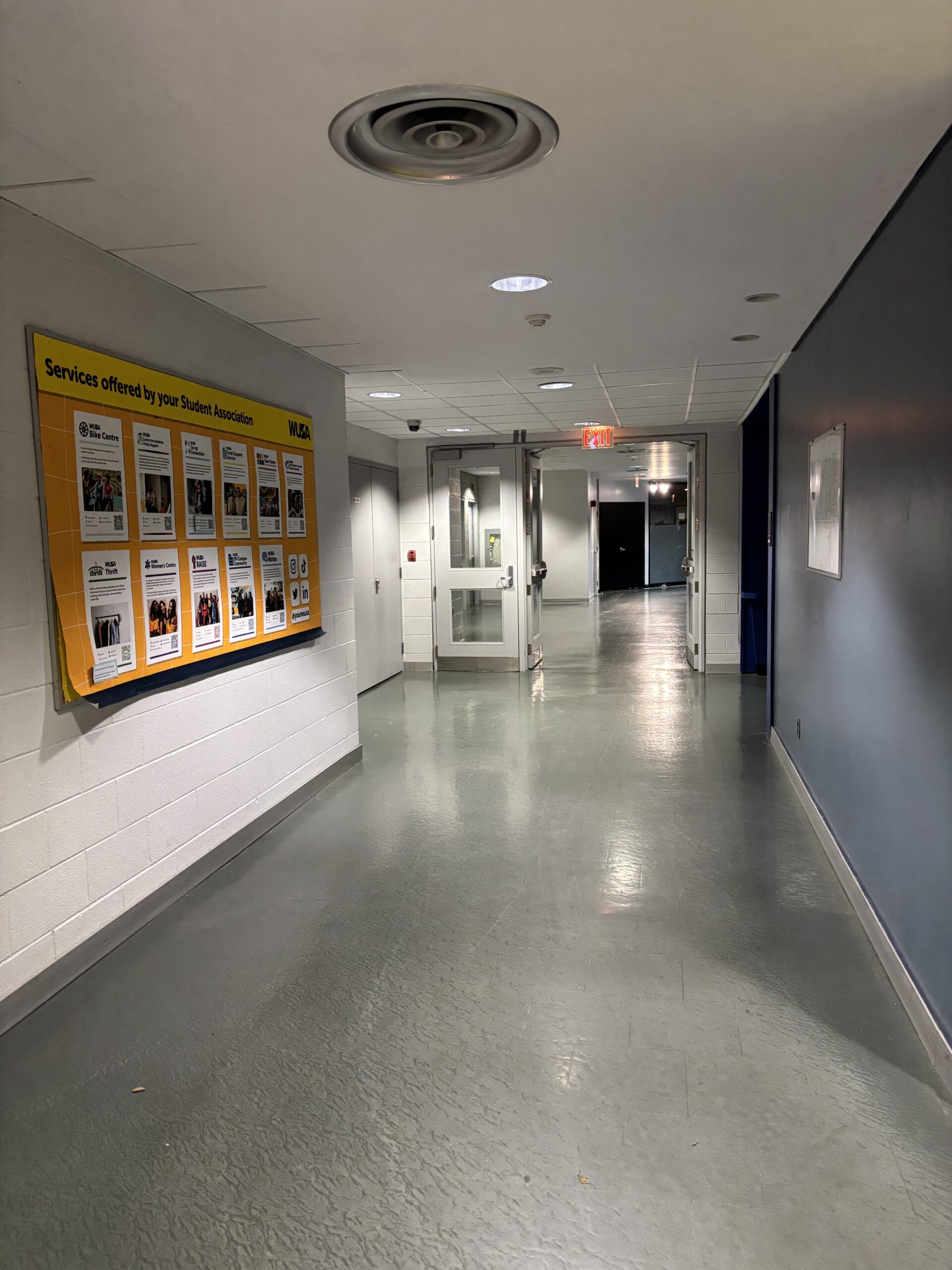 A hallway with a yellow bulletin board on the left wall leads to the glass doors of the Student Life Centre (SLC).