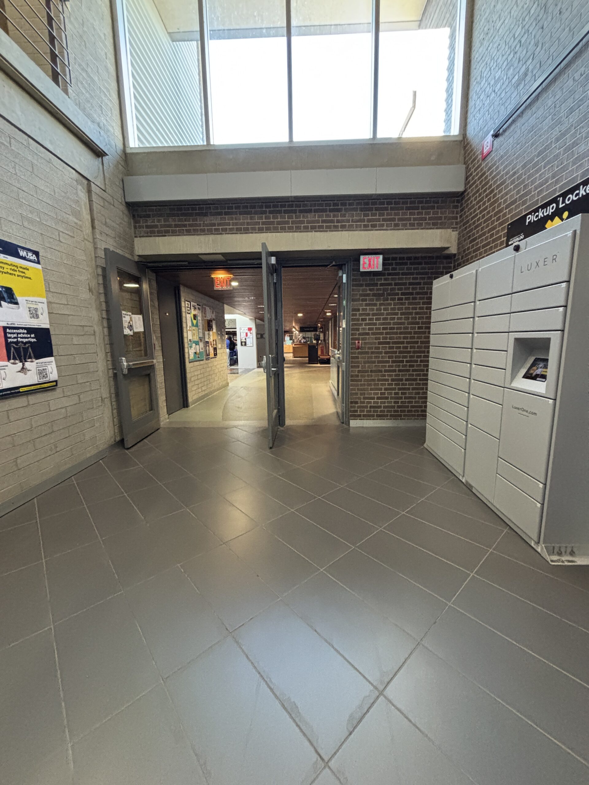 Wide hallway in SLC with double glass doors, a white parcel locker, and bulletin boards on brick walls.