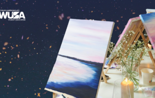 A canvas with a pastel landscape painting on an easel; flowers, candles, art supplies, and a Wine Not Paint glass set the scene for creativity.