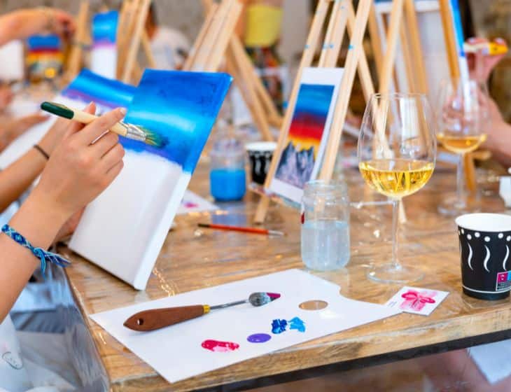 People at a Wine Not Paint event create colorful landscapes on canvases, surrounded by drinks, brushes, and art supplies.