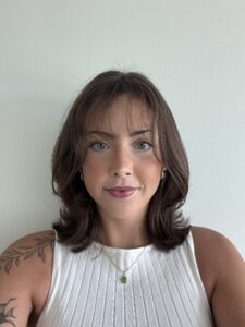 Young woman with short brown hair, wearing a white top and gold necklace, posing against a plain light background.