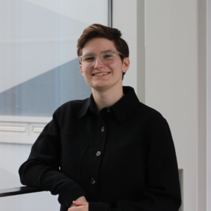 Person with short hair and glasses smiling, wearing a black jacket, standing indoors by a window.
