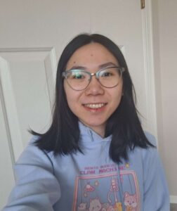 Woman with glasses and black hair smiles, proudly wearing a light blue hoodie with cartoon designs, standing indoors.