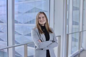 Woman in a gray blazer stands smiling with arms crossed in a modern glass building.