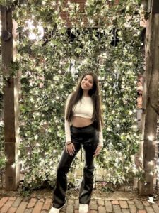 Woman smiling in front of a wall decorated with greenery and string lights on a brick pathway.