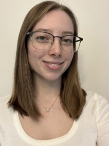 A young woman with straight brown hair, glasses, and a white top smiles at the camera against a plain background.