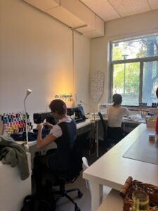 Two people work at sewing machines in a bright room with thread spools and a large window.