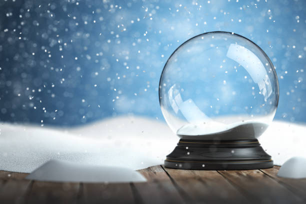 A snow globe with falling snow sits empty on a wooden surface against a snowy, blue background.