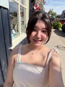 Woman in a light dress takes a selfie outside on a sunny day near a building with a Canadian flag.
