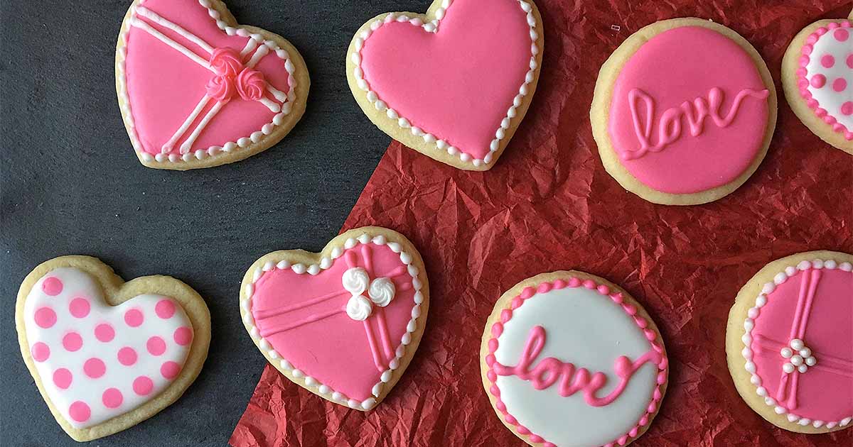 Heart and round sugar cookies decorated with pink, white, and red icing are arranged on red and black surfaces, perfect for a Valentine’s cookie decorating night.