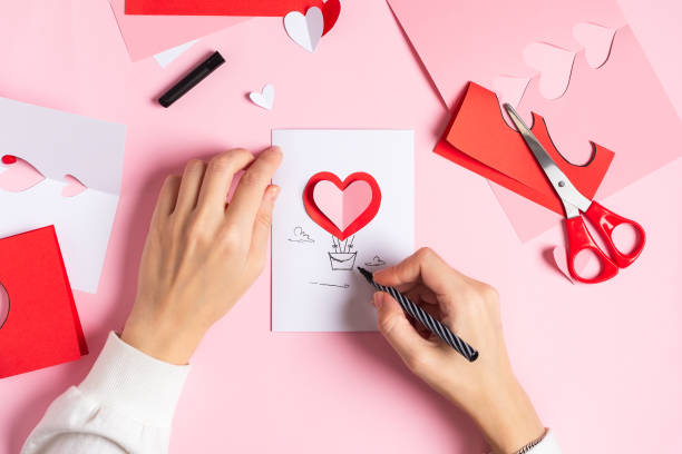 Hands making a handmade card with a red 3D heart on a pink table with paper, scissors, and pen—a perfect card making idea for Valentine’s Day.