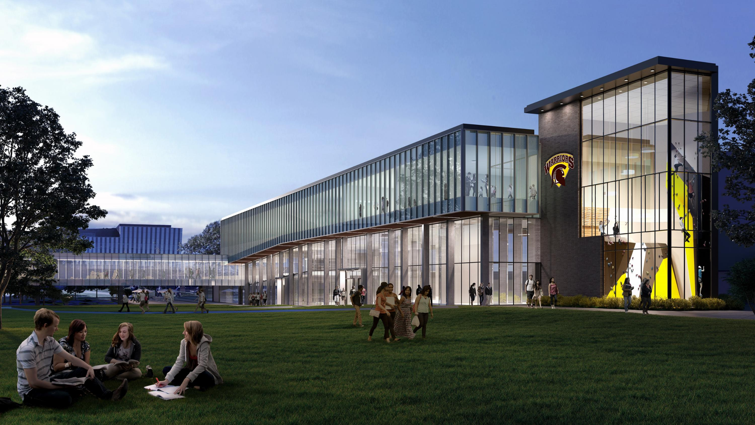 Students gather on a lawn in front of a modern glass building at dusk, with lights illuminating the interior.