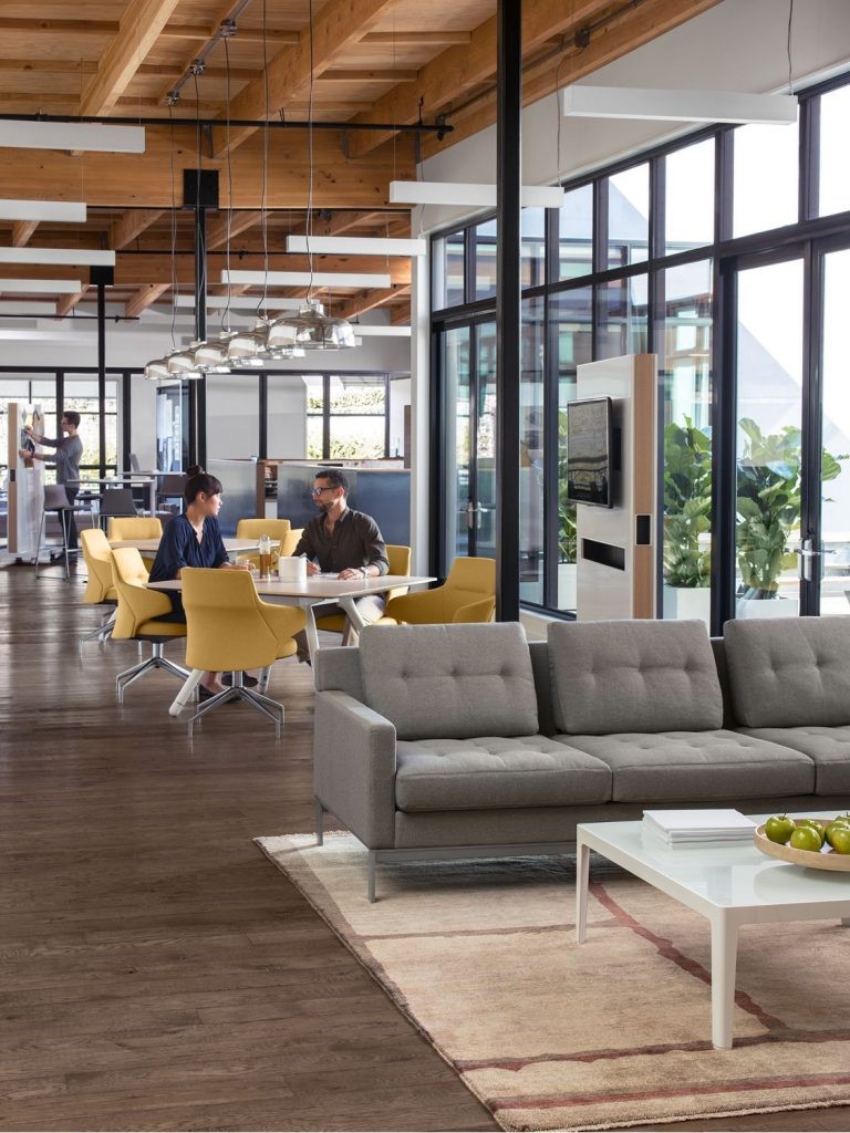 Modern open office with yellow chairs, glass walls, grey sofa, and people working and talking at tables.